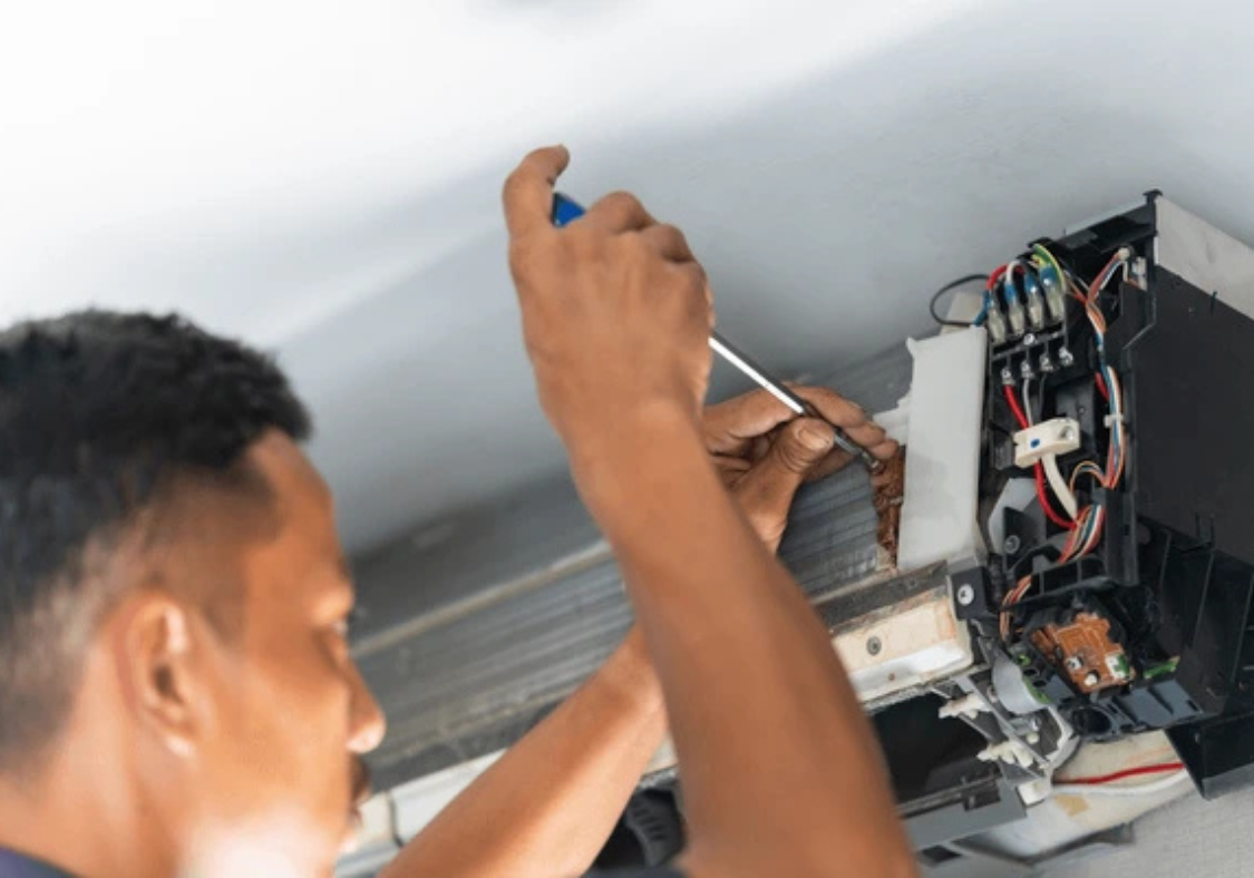 A man is repairing an air conditioner, using tools & focused on the unit's internal components.