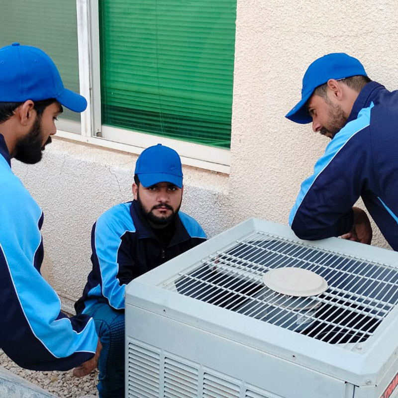 Three technicians in blue attire are working collaboratively on an air conditioning unit.