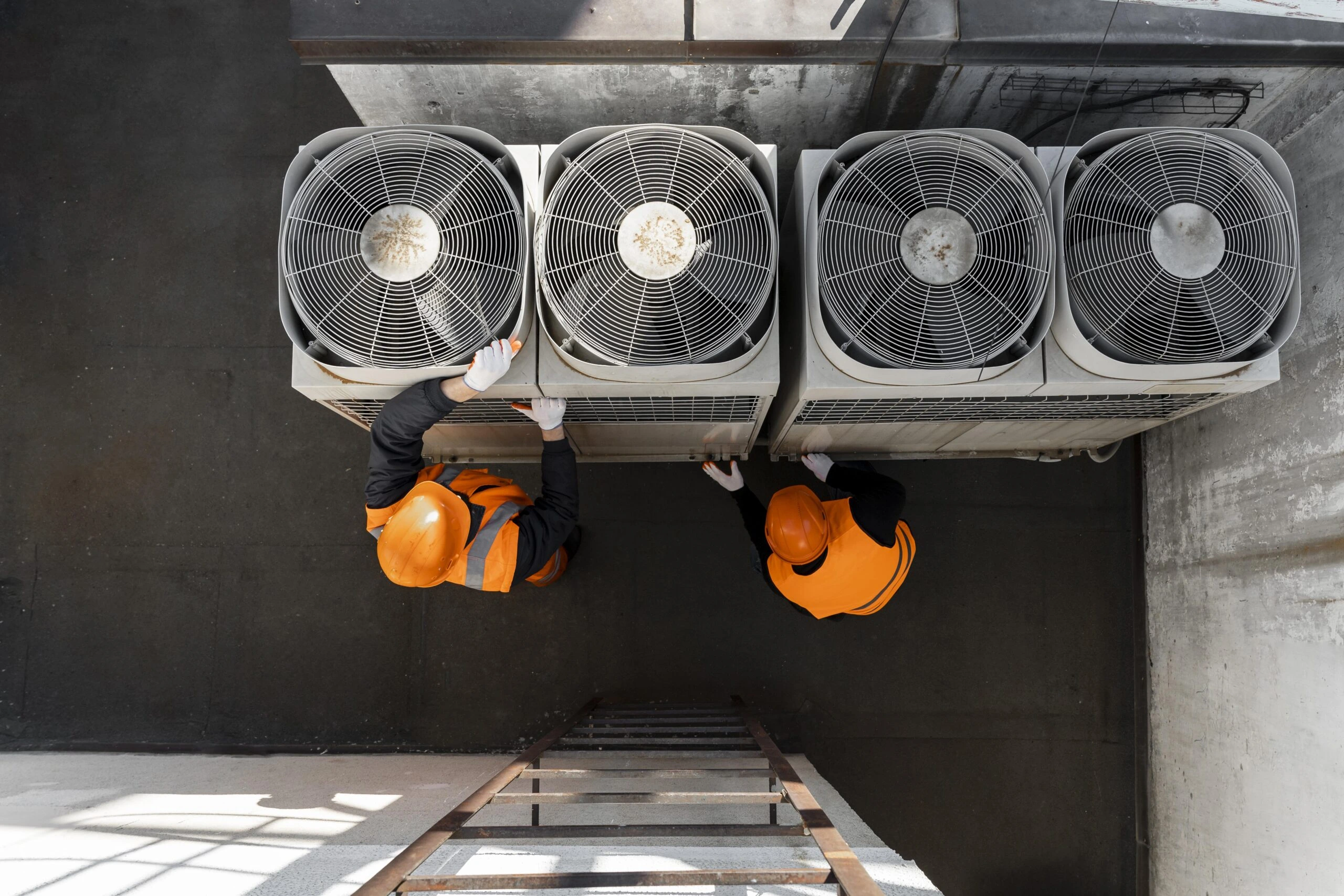 Two men in orange safety gear are repairing a large air conditioning unit on a rooftop.