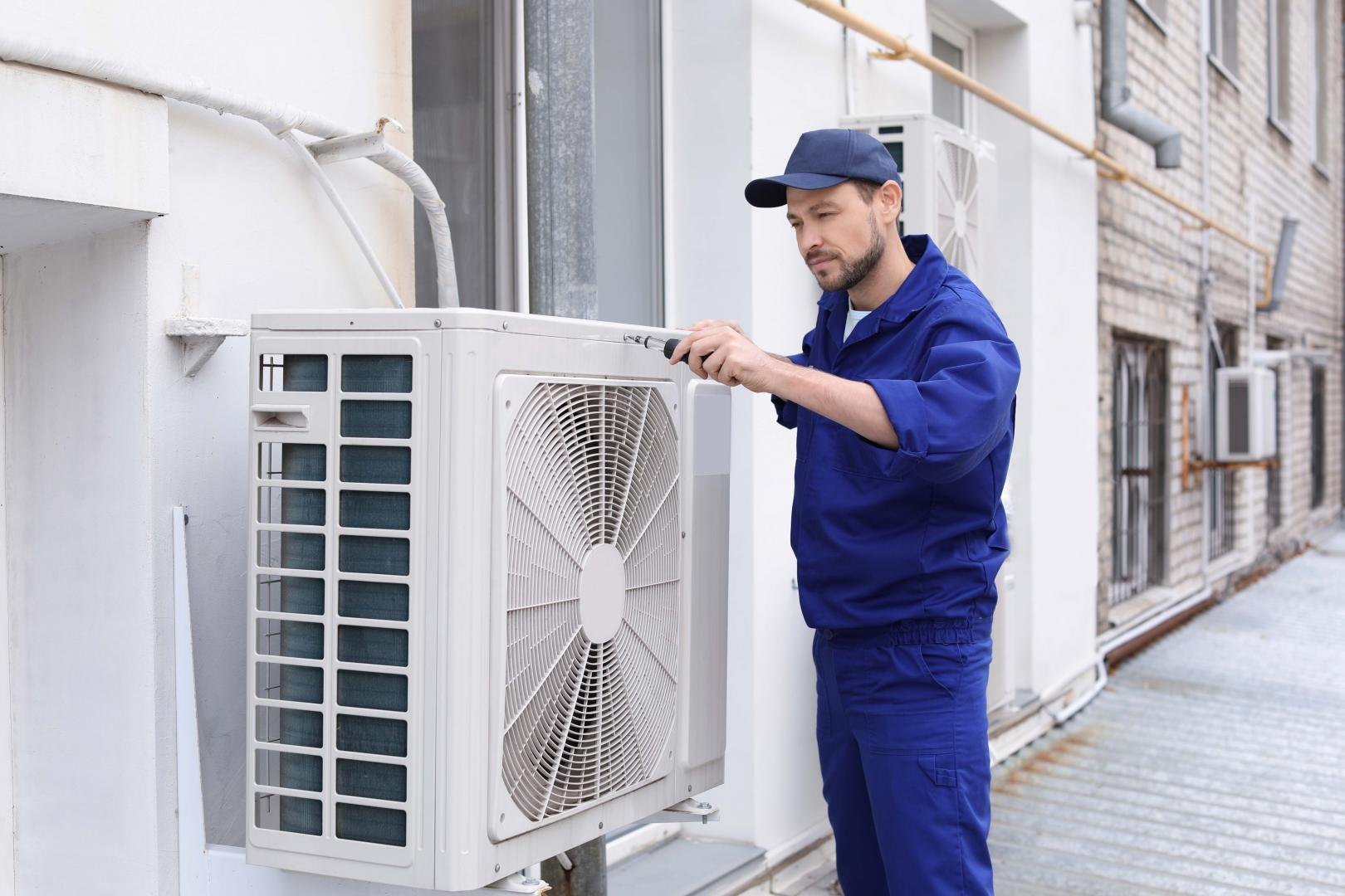 A man in a blue uniform repairs an air conditioner, focused on his task in a well-lit indoor setting.