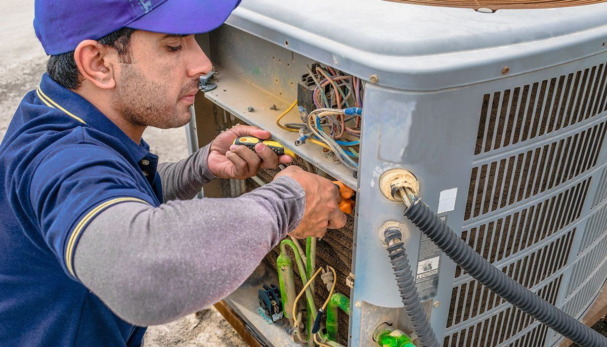 A man repairing an air conditioner, focused on the unit with tools in hand, in a well-lit indoor setting.