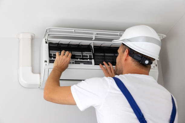 A man in a hard hat and overalls repairs an air conditioner, focused on his task in a residential setting.